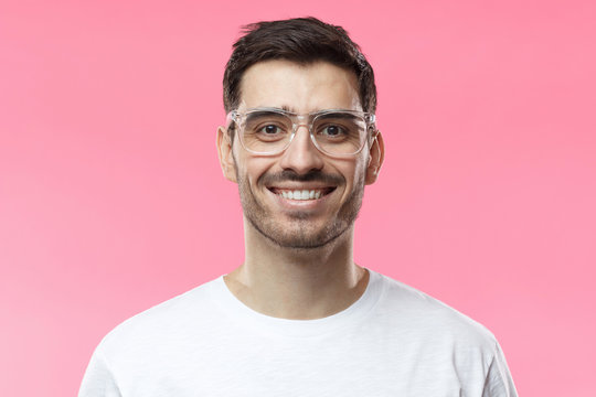 Close up shot of smiling attractive man in white t-shirt and trendy trasparent eyeglasses isolated on pink background