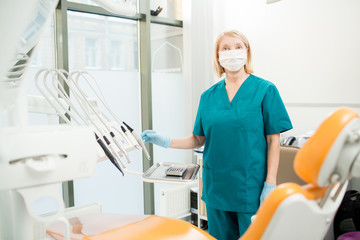 Nurse in uniform, mask and gloves looking at camera while standing by workplace of dentist