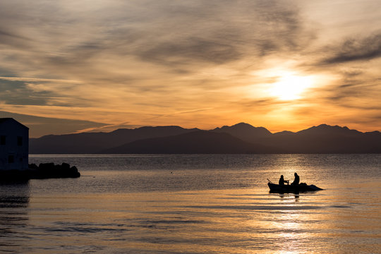 Silhouettes of two men rowing in a small fishing boat at a colorful golden cloudy sunrise in Corfu, Greece. Horizontal image