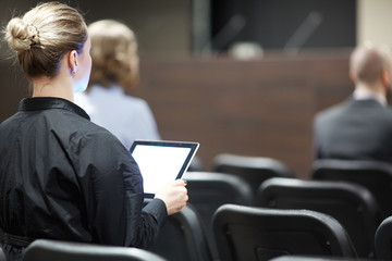 One of participants of conference with tablet sitting in row of chairs and listening to speaker