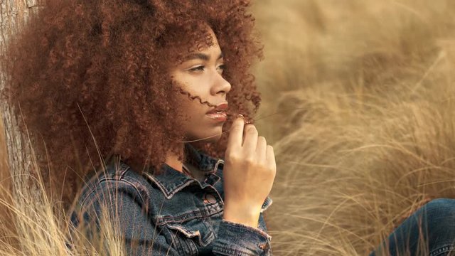 black mixed race woman with big afro curly hair sits on lawn with high grass on sunset. Denim look on hay