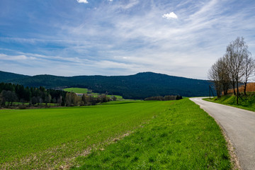 Straße in der Landschaft im Bayerischen Wald mit Wiese und Berge im Hintergrund und Wolken am Himmel