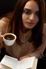 A young beautiful girl in a warm sweater holds a cup of coffee and reads a book, a cute portrait