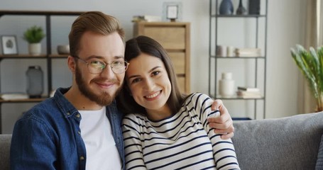Portrait shot of the happy attractive smiled just married couple looking at each other and smiling to the camera while sitting and embracing on the sofa at home. Indoor - Powered by Adobe