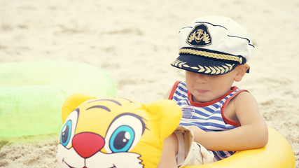 Little Caucasian boy in sailor suit and cap sitting on the beach in swim ring and laughs.