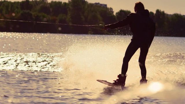 Wakeboarder surfing on river behind motorboat. Extreme wakeboarding at sunset. Wakeboard rider moving fast in splashes of water at golden sunset. Extreme water sport
