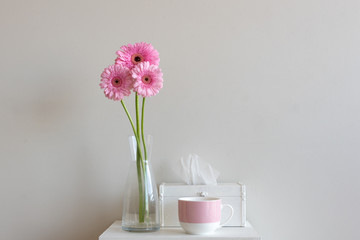 Tall pink gerberas in glass vase with tissue box and pink mug on small white table against neutral wall background