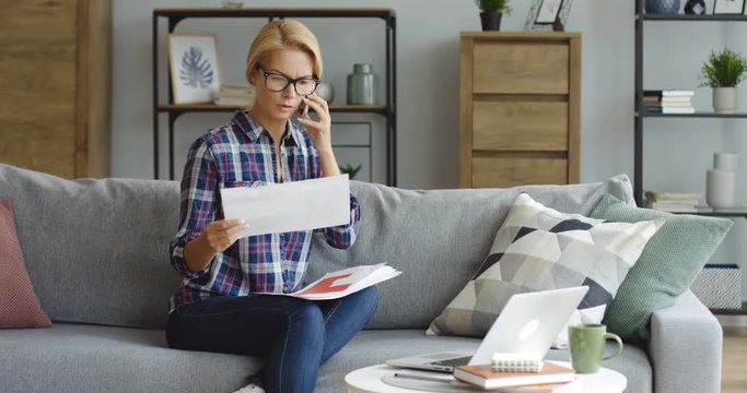 Pretty blonde woman in glasses and in motley shirt sitting on the sofa, speaking on the phone and cheking documents at home. Inside