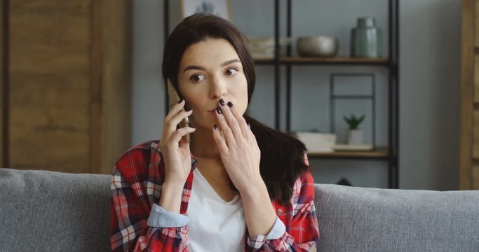 Close up of the attractive brunette young woman in the red plaid shirt talking on the smartphone and getting to know shoking news at home. Indoor