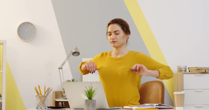 Young good looking Caucasian woman in a yellow sweater working at the table with a laptop computer on it in the cozy office and then stretching her back and neck. Indoor