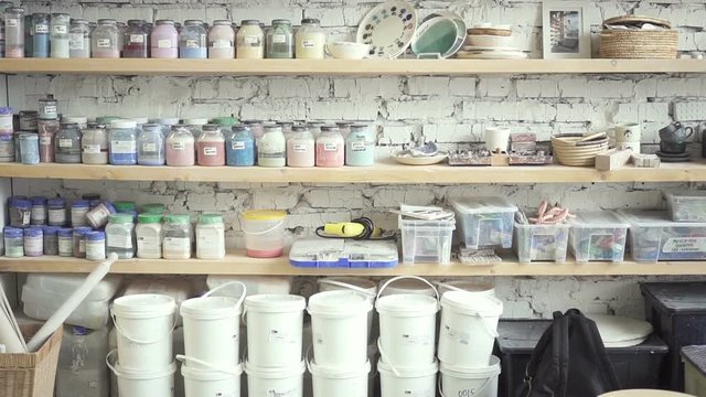 Jars with colors stand on shelves in art workshop indoors. Paints for painting and ceramics products is on wooden racks in modern studio. In bright room with white brick walls, there are many objects