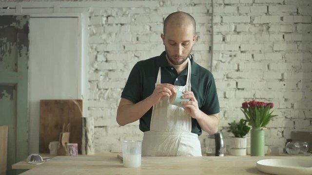 In workshop man processes hand-made article of clay with calcination solution. Master in an apron applies a protective layer to surface of dishes for further processing of ceramics.