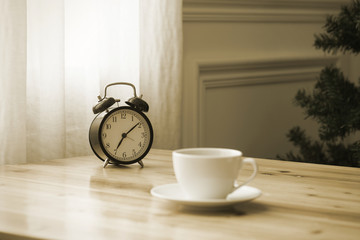 A cup of coffee and a pot of plants on a wooden table by the window. Focus on clock.