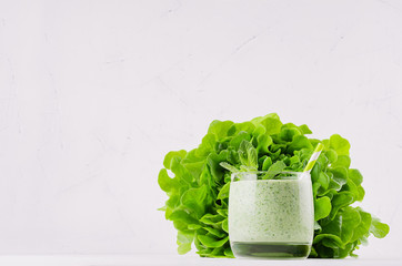 Green vegetable smoothie in glass with straw, mint twig, lettuce, copy space. Soft white wooden board background.