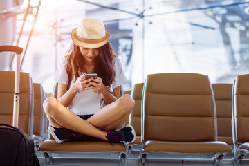 Asian woman teenager using smartphone at airport terminal sitting with luggage suitcase and...