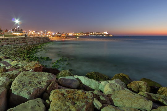 Nocny widok zatoki Morza Śr&oacute;dziemnego, umocnionego brzegu i nocnych świateł w Tel Awiwie, Izrael, w oddali widoczna rozświetlona Jaffa