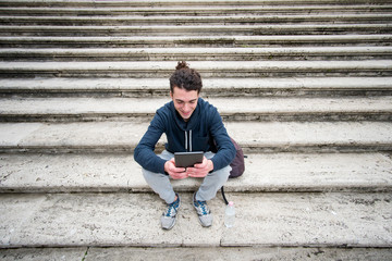 Handsome young man in sports outfit with curly hair sitting on stairs and working on tablet