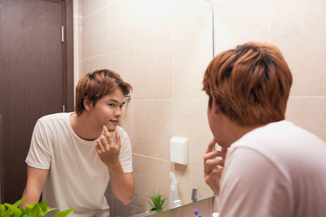 Young asian man looking in mirror and checking his skin in bathroom