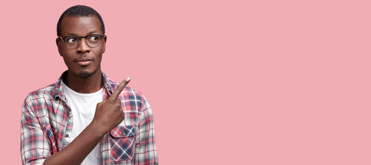 Attractive young male employee with dark skin, wears spectacles and checkered shirt, indicates with...