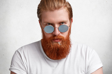 Portrait of confident brutal male with stylish haircut, has thick long fuzzy red beard and mustache, wears round shades and white casual t shirt, poses alone against concrete wall. Fashionable hipster