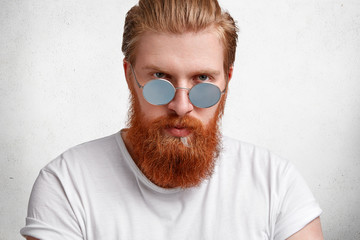 Brave handsome man with attractive serious look, has stylish red beard and mustache, wears sunglasses, dressed casually, isolated over white concrete background. Fashionable male boss models indoor