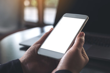 Mockup image of hand holding white mobile phone with blank desktop screen and laptop on table