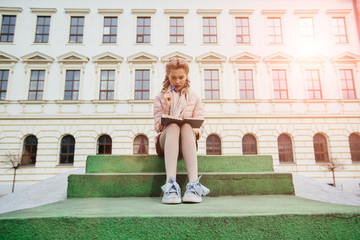 Full height of european female student thinking and writing something in notepad while sitting on the stairs over university building background. Sun glare effect.
