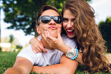 Selfie-portrait of funny couple  lying on grass in summer park. Girl with long curly hair, red lips...