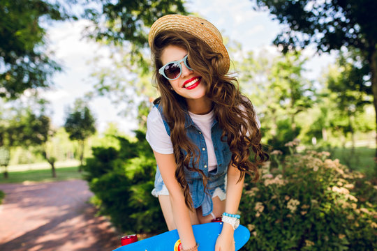 Portrait of attractive girl with long curly hair in hat posing with skateboard in summer park. She wears jeans jerkin, sunglasses. She is smiling.