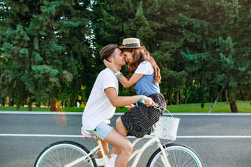 Gentle couple in love on bike on road on forest background. Handsome guy in white T-shirt is driving a bike, girl with long curly hair in hat sitting on the handlebars and hugs him.