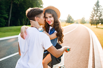Romantic couple in love on bike on road on sunset. View from back of handsome guy in white T-shirt  driving a bike, girl with long curly hair in hat, hugs him and smile to camera with red lips