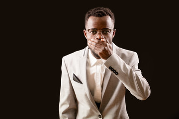 Young scared Afro man in glasses keeping his mouth closed by his hand on dark background. open-eyed...