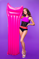 Fototapeta na wymiar Full-length photo of attractive girl with long curly hair posing to camera on purple background in studio. She wears swimsuit and holds pink air mattress near.
