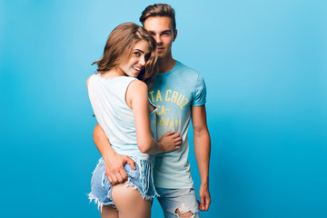 Young couple is hugging on blue background in studio. Handsome guy pulls up shorts of pretty girl with long curly hair.