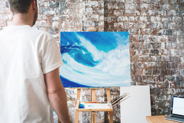 Artist stands in front of the easel with a canvas and considers his work. Painter in art studio
