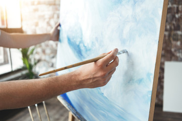 Male hand of artist closeup holding paintbrush on background of canvas on easel. Painter drawing ocean wave in loft studio. Flare effect. Hobby concept