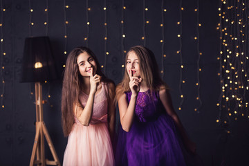Merry girlfriend. Two girls in pink and purple dresses show shh, in studio on dark background. Copy space
