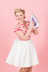 Young pin-up styled girl holds iron on pink background. Beautiful pin-up blonde woman with curls prepare iron for work isolated in studio. Blonde lady happy to have iron in hands. Pretty blonde girl