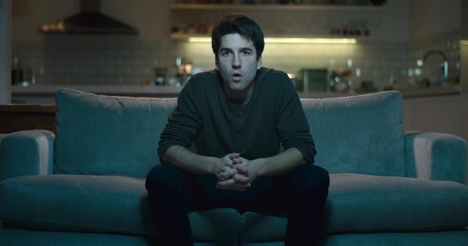 Man sitting on the edge of a sofa amazed and astonished by what he is watching.