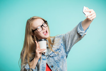 Young cheerful woman taking selfie on phone on blue background. The girl is wearing jeans and holds...