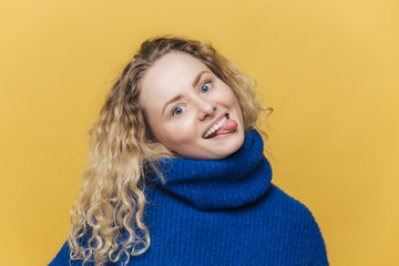 Funny comic curly young woman with positive expression, shows tongue, has fun with friends, wears blue sweater, makes grimace, isolated over yellow studio background. People and fun concept.