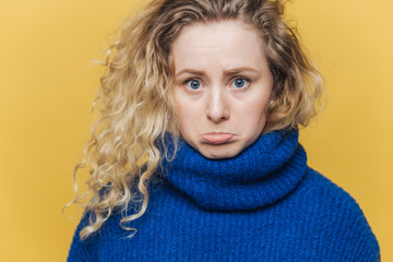 Attractive offended young female has curly bushy hair, curves lower lip in dissatisfaction, feels upset after quarrel with husband, wears blue sweater, isolated over yellow studio background.