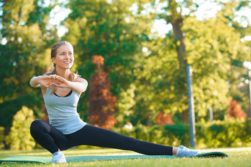Frontview of young pretty girl doing stretching on the park's grass. Wearing sport clothes in vlack...