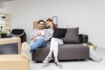 Young couple sitting in new empty room with unpacked boxes and looking at laptop while he drink cofe from mug