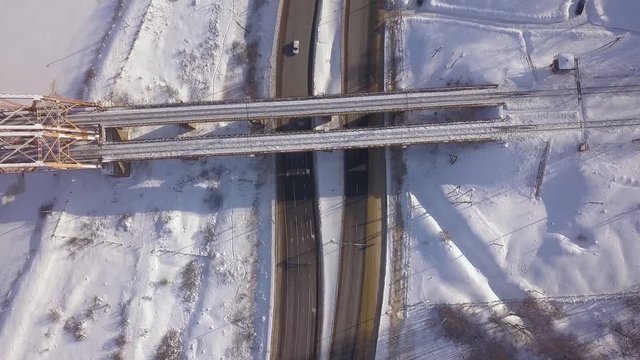 Cars driving on winter highway under suspension railway bridge drone view. Car traffic on snowy road on winter landscape. Aerial view train bridge over highway road.