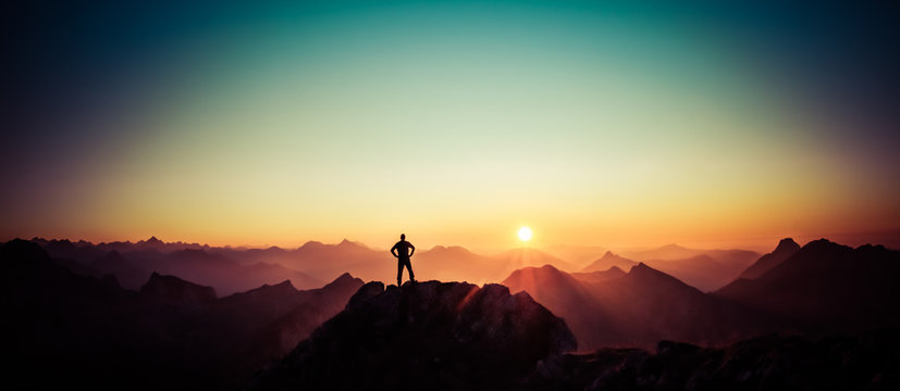 Man reaching summit enjoying freedom and looking towards mountains sunrise.