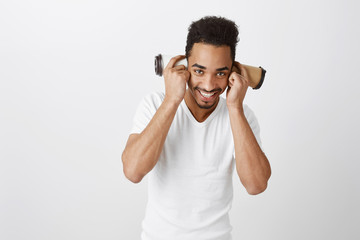 Fototapeta premium Coffee during day takes sleep away. Funny emotive african-american model with afro hairstyle and white t-shirt, smiling broadly, being in good mood, holding cups over ears, standing against gray wall