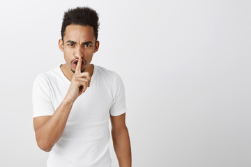 Stop talking. Studio shot of outraged bothered dark-skinned guy with afro haircut and tattoo, frowning, making shush motion with disdain and anger, holding index finger over mouth