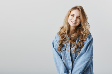 Sister takes care of infant while parent at work. Charming tender feminine woman with blond hair, holding hands together in front of chest, wearing denim jacket and smiling friendly at camera