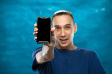 Positive blurred young male hipster points finger at black screen smartphone on blue background. Place for text